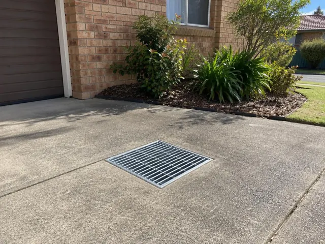Concrete driveway with a seated stormwater grate beside a brick house and garden in Old Toongabbie